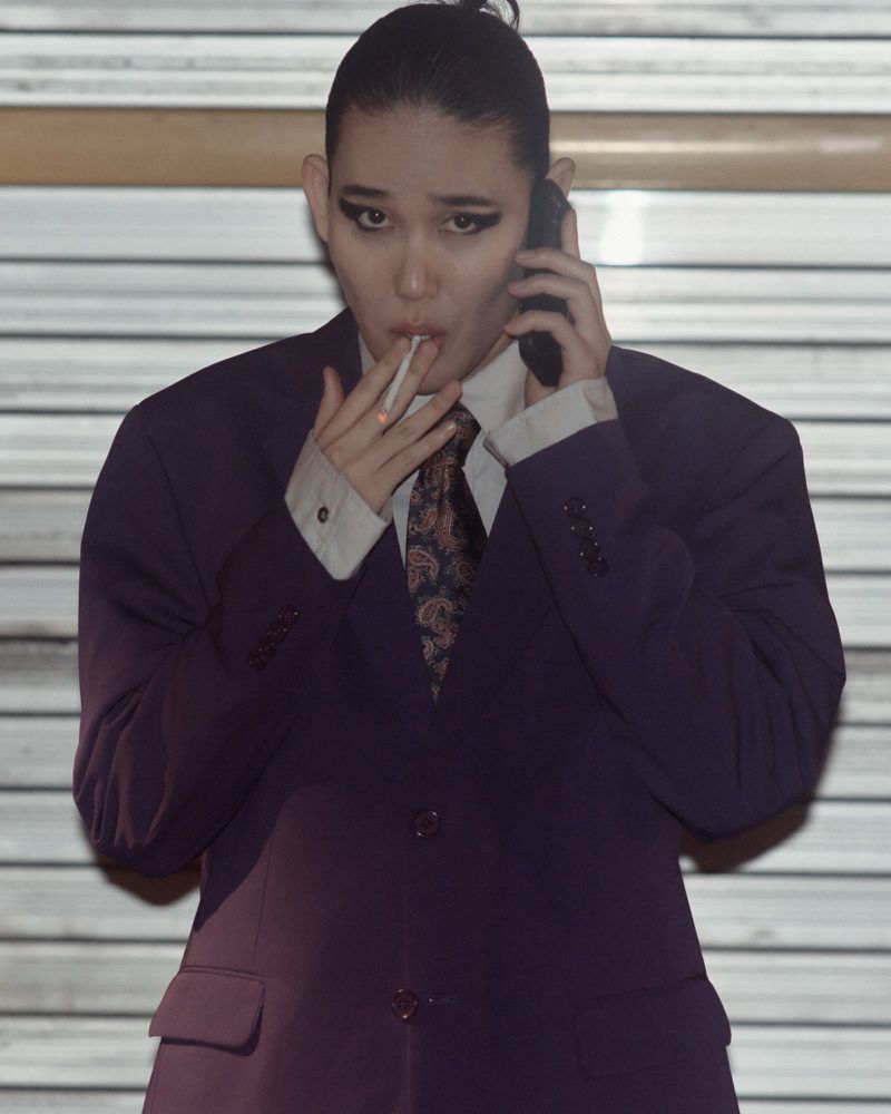 A person wearing a suit is talking on a smartphone while holding a cigarette in an indoor environment with a neutral color palette.