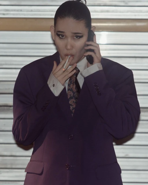 A person wearing a suit is talking on a smartphone while holding a cigarette in an indoor environment with a neutral color palette.