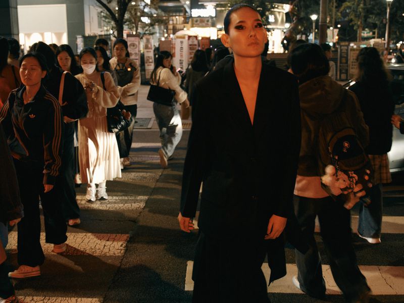 Woman in black coat crosses a crosswalk at night surrounded by a crowd of pedestrians in an urban setting with street lights.