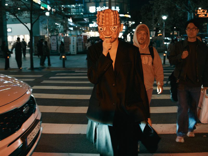 Model wearing a sculpted orange traditional mask and an oversized black blazer walking across a night crosswalk.