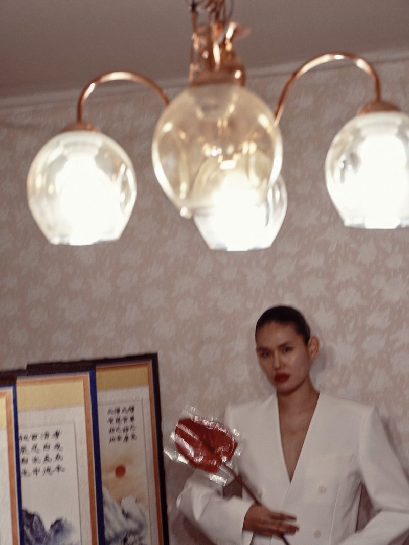 Elegant woman in a white suit poses indoors, holding a red object, with decorative paintings and a chandelier adding to the ambiance.