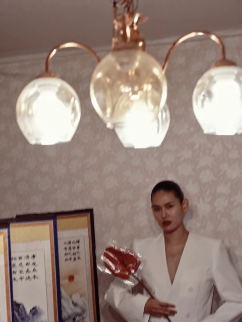 Elegant woman in a white suit poses indoors, holding a red object, with decorative paintings and a chandelier adding to the ambiance.