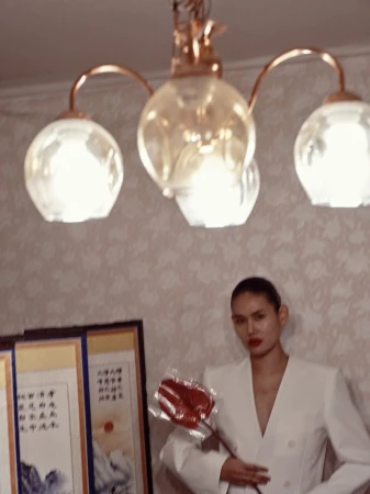 Elegant woman in a white suit poses indoors, holding a red object, with decorative paintings and a chandelier adding to the ambiance.