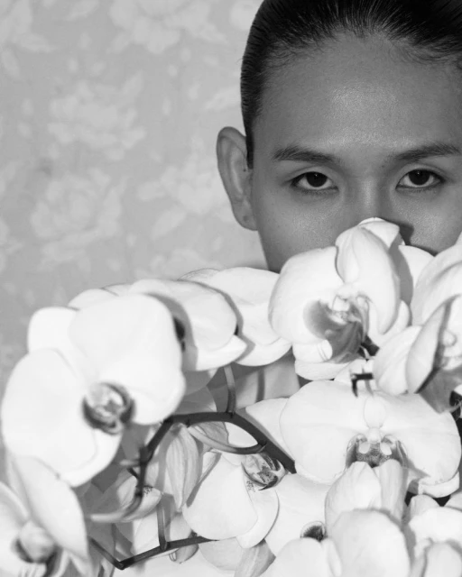 Close-up black and white portrait of a young adult peering over a bouquet of white orchids with a floral wallpaper background.