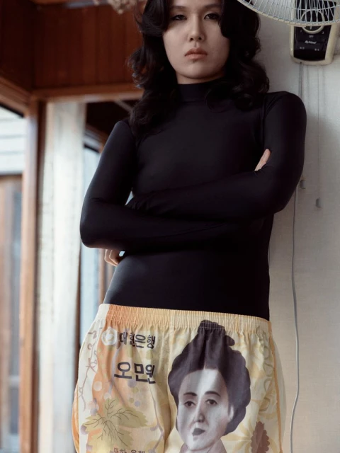 Woman in long-sleeved shirt and money-themed shorts stands in an indoor setting with a vintage fan, creating an editorial-style portrait.