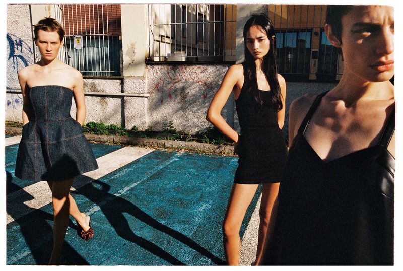 Three women model strapless and spaghetti-strap dresses on a city street with blue pavement, graffiti, and a barred window.