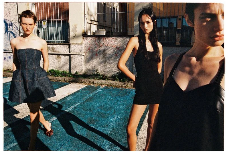 Three women model strapless and spaghetti-strap dresses on a city street with blue pavement, graffiti, and a barred window.