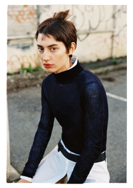 Portrait of a woman with short dark hair wearing a navy turtleneck sweater and white skirt against an urban background with graffiti.