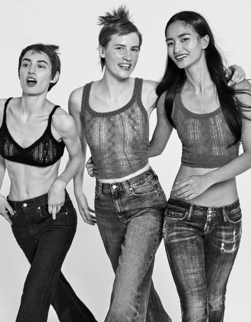 Three young women in jeans and knit tops pose in a studio. One woman has her hand on another's shoulder in a black and white shot.