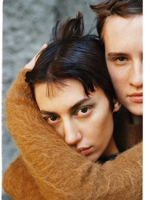 Portrait of a couple embracing, woman with olive skin and short dark hair wearing a furry brown sweater and a muted grey background.