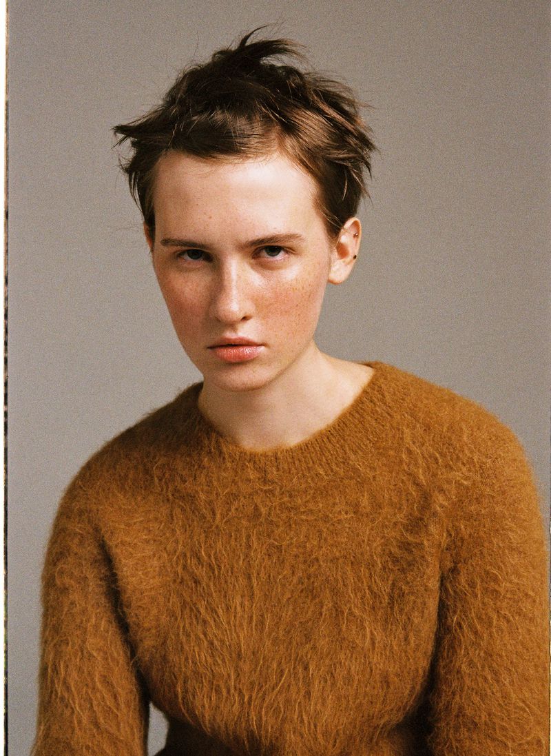 Portrait of a fair-skinned young adult female with short brown hair and a fuzzy brown sweater, posing indoors against a neutral grey background.