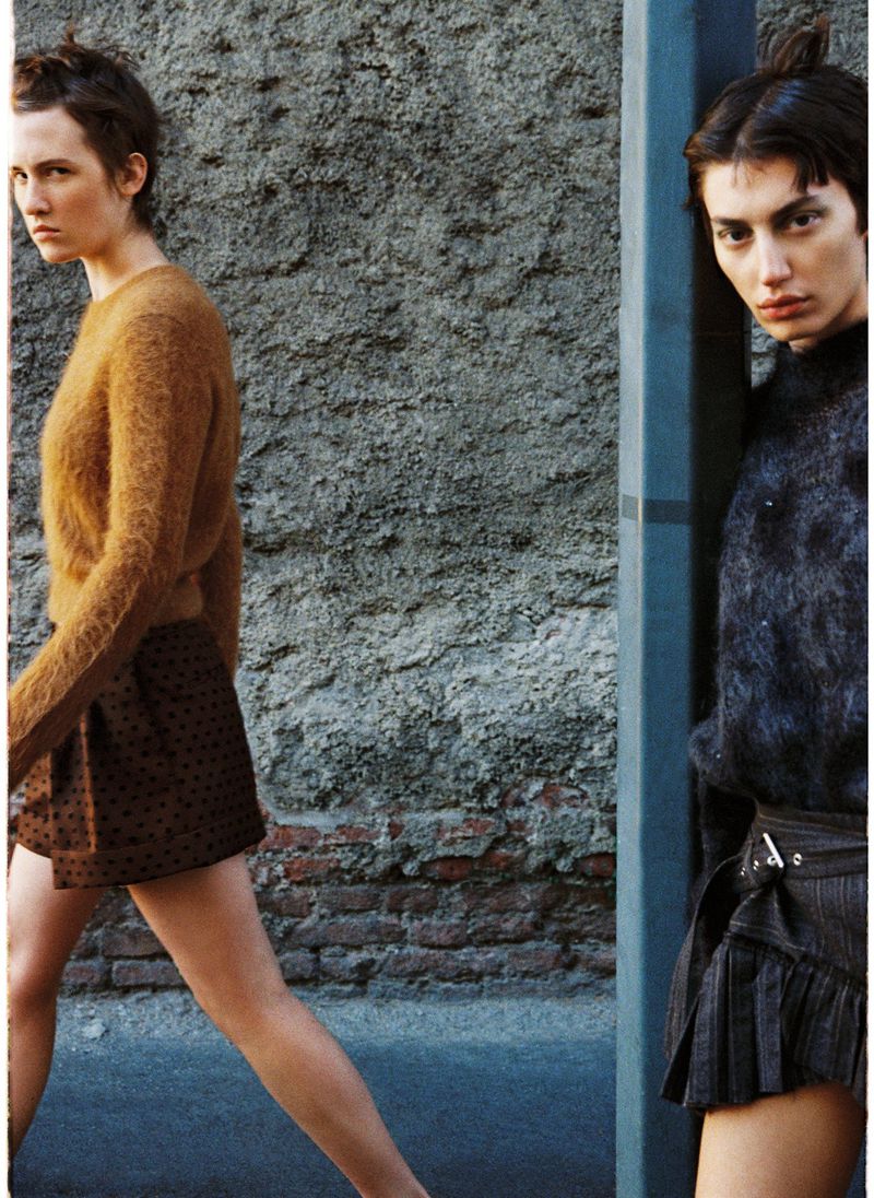 Two women in fashionable attire pose against a textured urban wall, one in a brown sweater, the other in a dark top with a short pleated skirt.