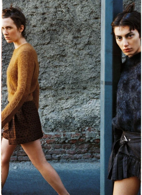 Two women in fashionable attire pose against a textured urban wall, one in a brown sweater, the other in a dark top with a short pleated skirt.