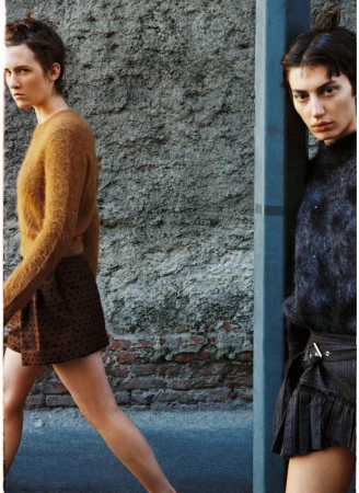 Two women in fashionable attire pose against a textured urban wall, one in a brown sweater, the other in a dark top with a short pleated skirt.