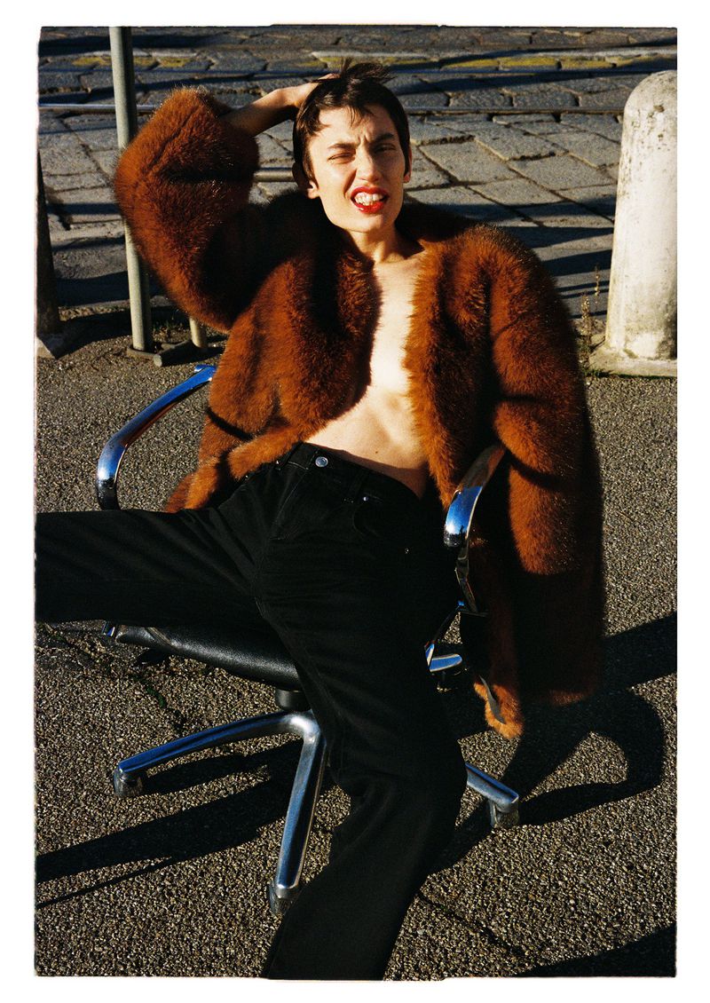 A woman in a fur coat sits on a metal and leather chair outdoors on a stone surface. Her head rests in her hand, looking into the camera.