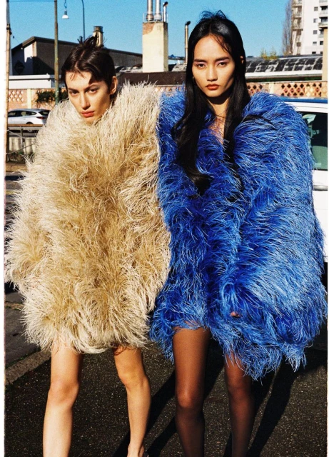 Two women wearing large, colorful, textured coats stand on a city street in an outdoor fashion shot with buildings and sky in the background.