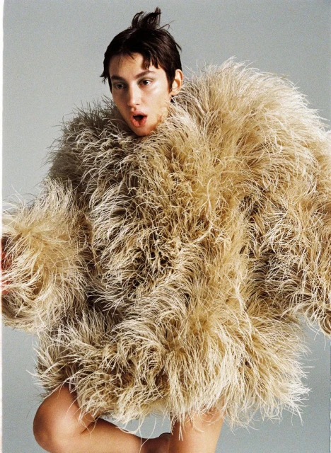 Adult female posing in an oversized feathery tan jacket against a gray background, looking at the camera with an open mouth. 