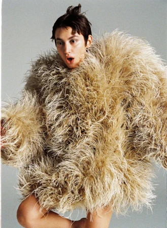Adult female posing in an oversized feathery tan jacket against a gray background, looking at the camera with an open mouth. 