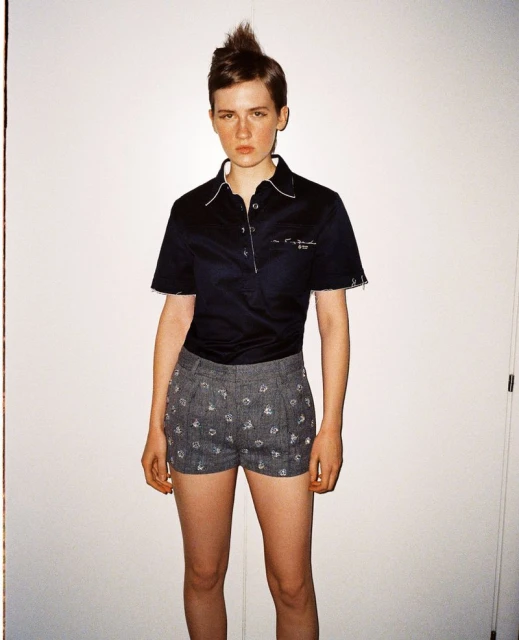 Full-length studio shot of a young adult woman with a mohawk hairstyle wearing a black collared shirt and patterned gray shorts.