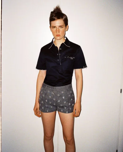 Model with short spiky hair wearing a dark short-sleeve polo shirt and patterned grey shorts against a plain white backdrop.