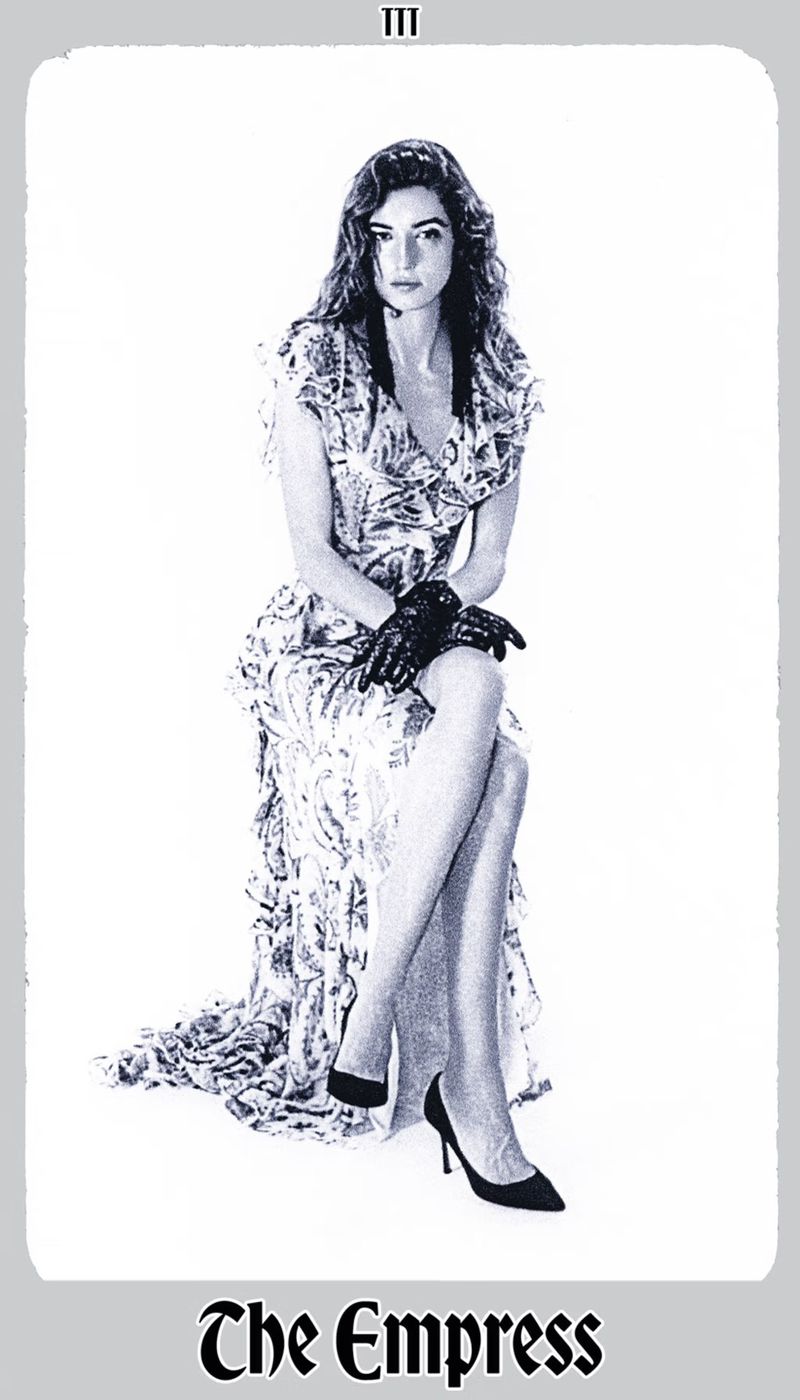 Monochrome portrait of a woman with long hair in a floral dress and lace gloves, seated in a studio setting, reminiscent of tarot card art.