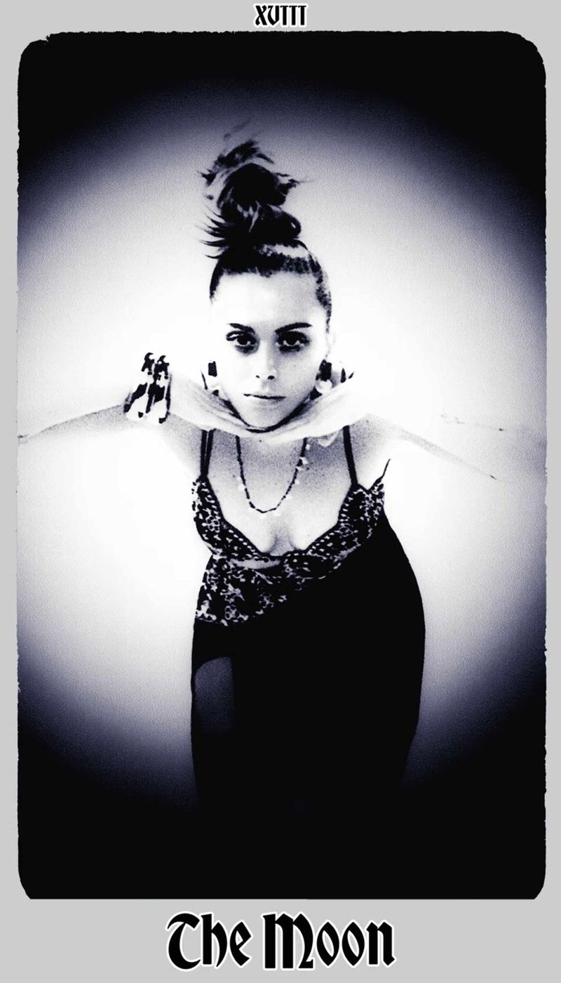 Monochrome tarot card image featuring a woman with a high bun, adorned with necklaces, posing with outstretched arms in a studio setting.