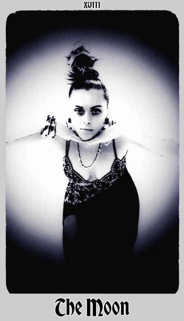 Monochrome tarot card image featuring a woman with a high bun, adorned with necklaces, posing with outstretched arms in a studio setting.