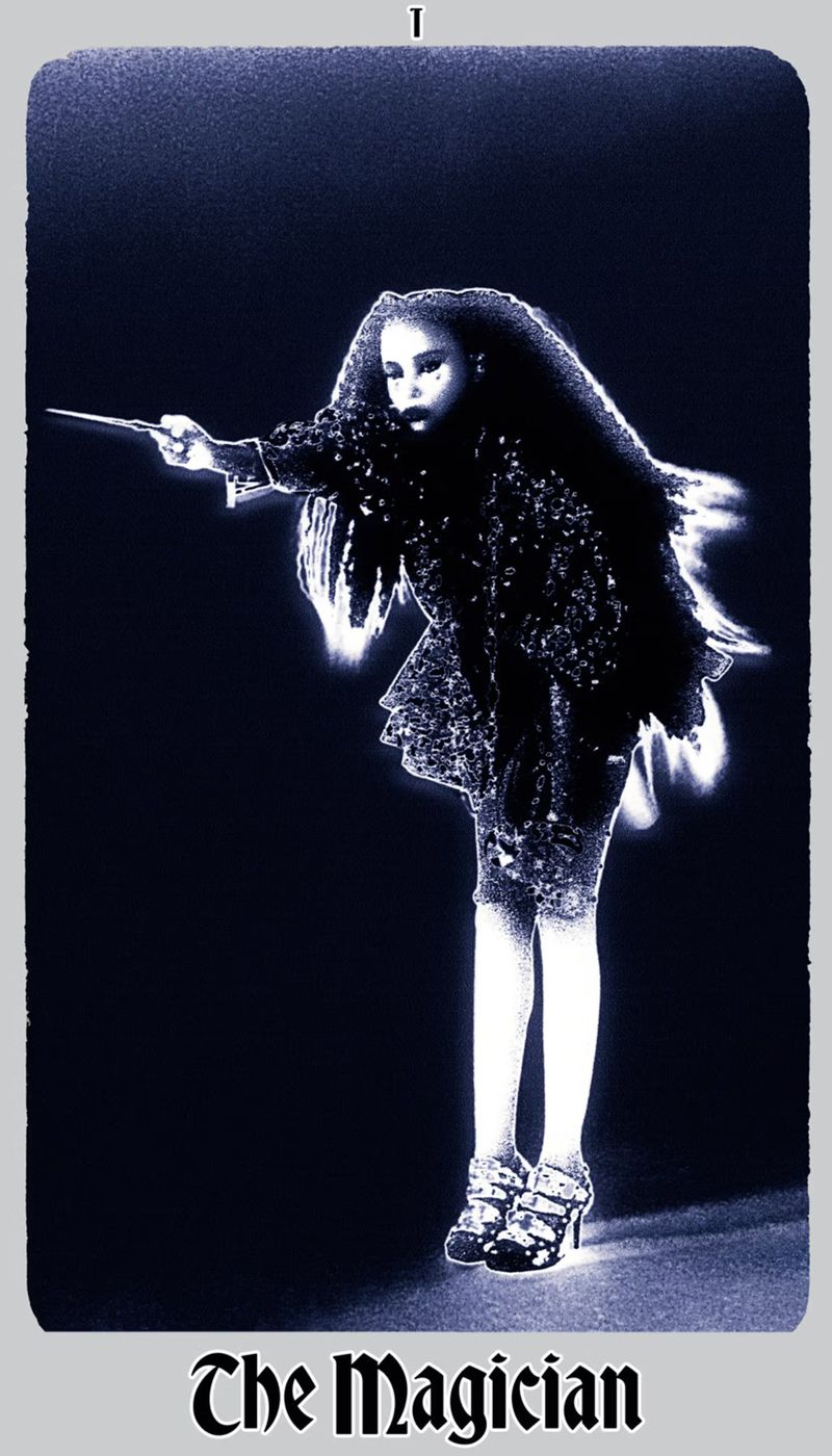 A woman in an embellished dark outfit stands in a dark space, holding a stick out. The image has a blue tint and is framed by a border.