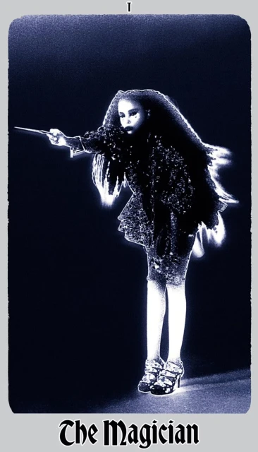 A woman in an embellished dark outfit stands in a dark space, holding a stick out. The image has a blue tint and is framed by a border.