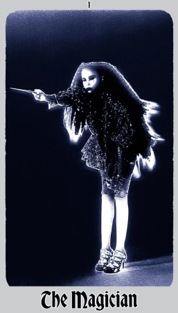 A woman in an embellished dark outfit stands in a dark space, holding a stick out. The image has a blue tint and is framed by a border.