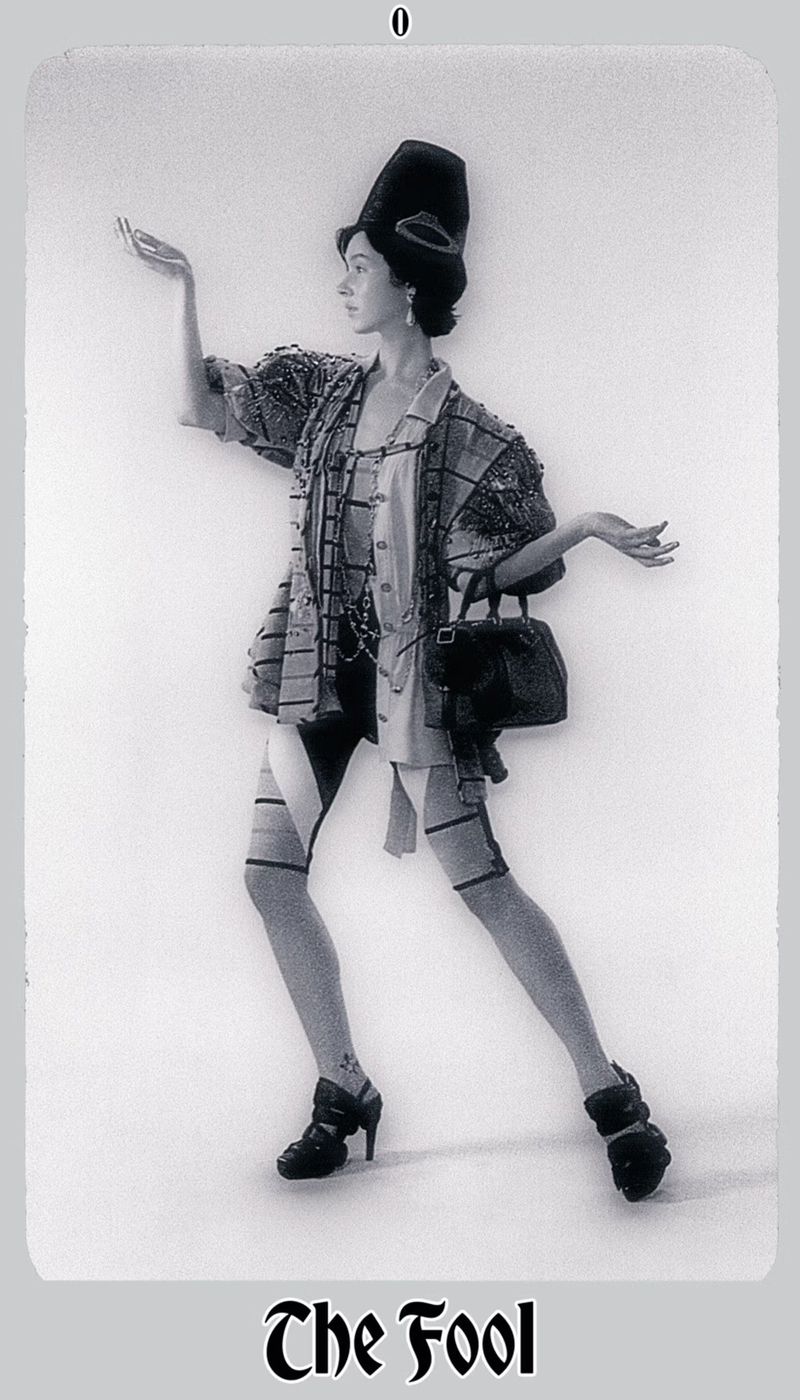 A female fashion model poses indoors, wearing an avant-garde outfit with a jacket, dress, leggings, heels, handbag, and a unique tall hat.