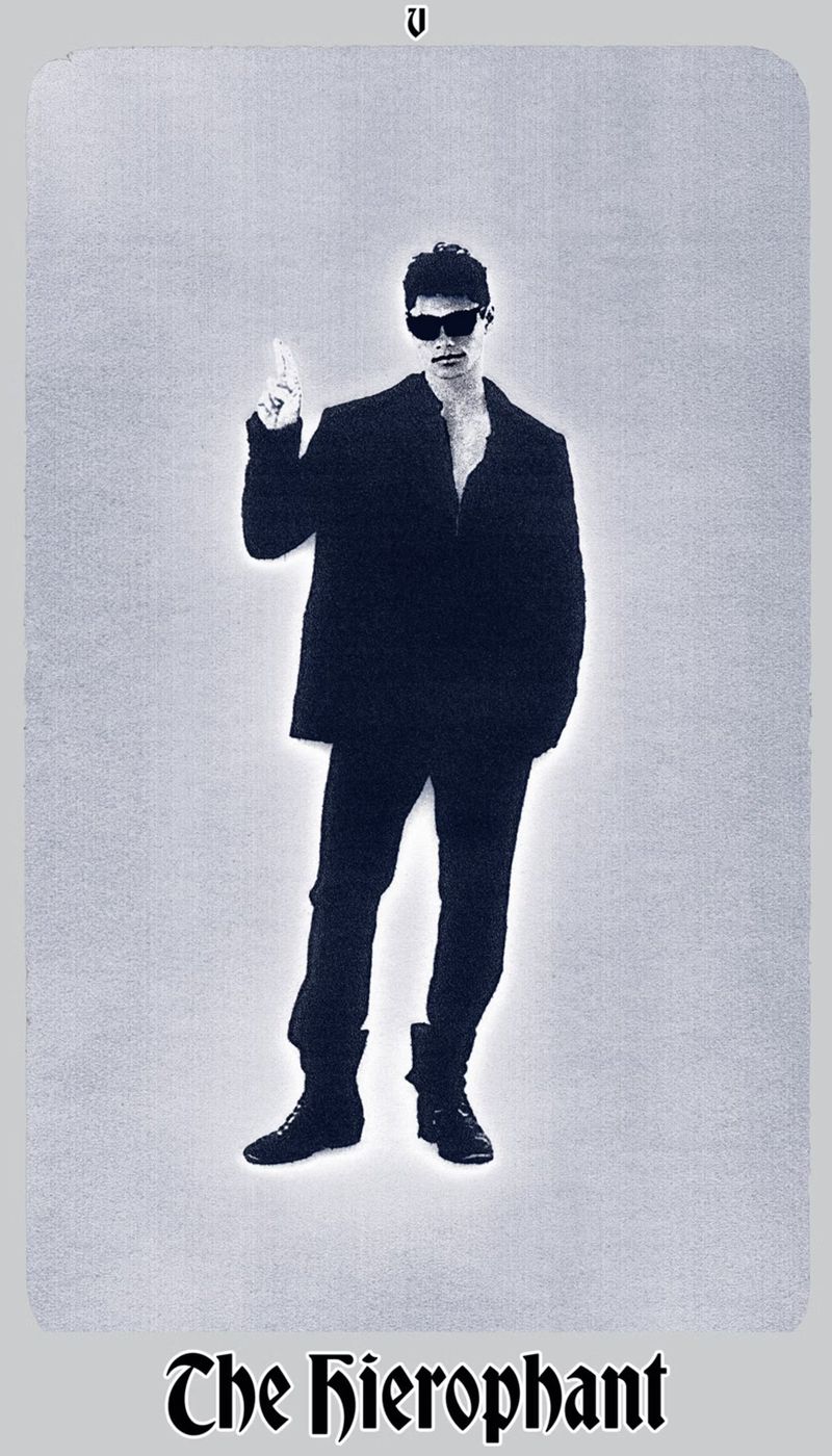 Person in dark suit and sunglasses stands in front of a light grey background, raising one hand with a finger pointed up.