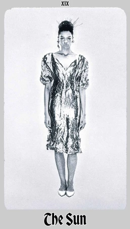 Full-length studio shot of a woman in a shimmering silver dress, with hair styled up and white shoes, against a white background.
