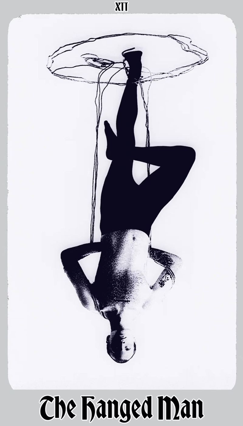Black and white art of a nude man hanging upside down suspended from above, an artistic rendering with visible text.