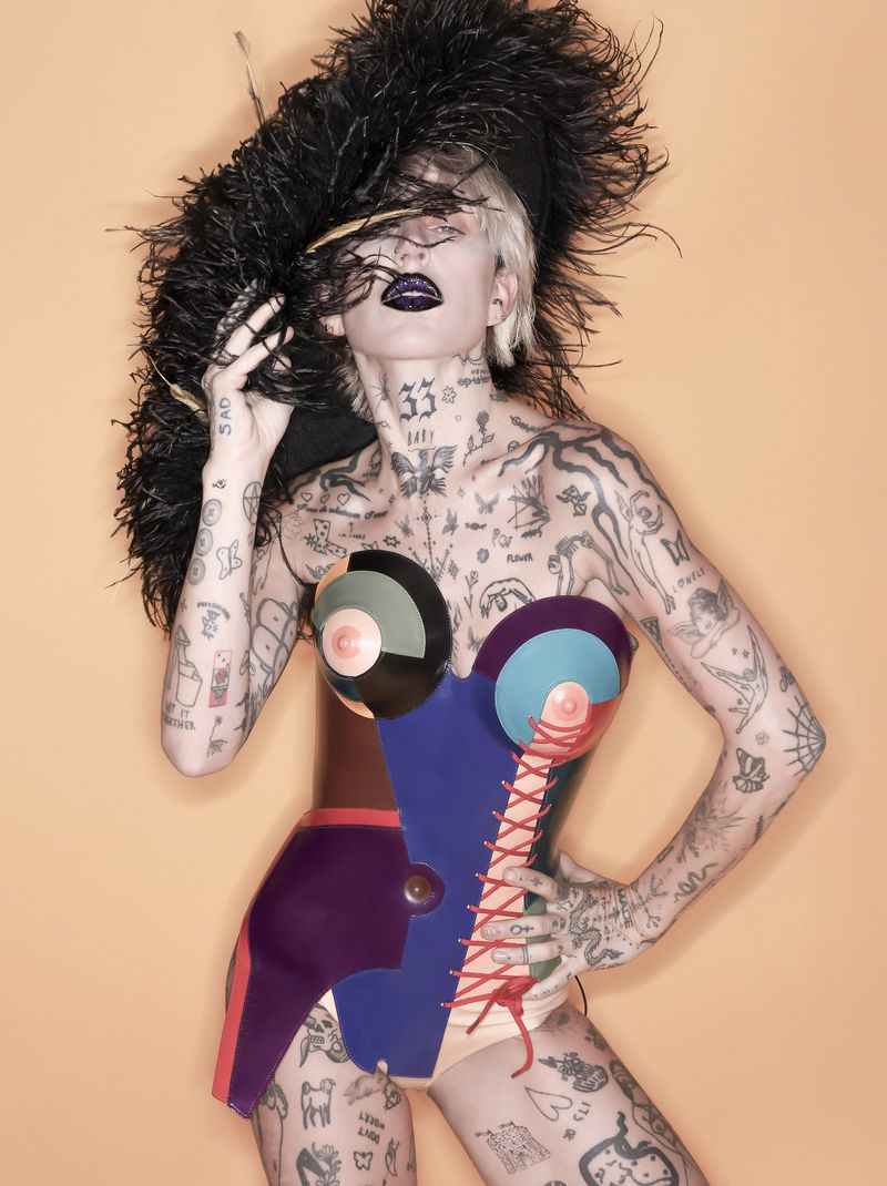 Woman with tattoos and an avant-garde outfit poses in a studio setting, holding a large feathered accessory. Studio lighting is even.
