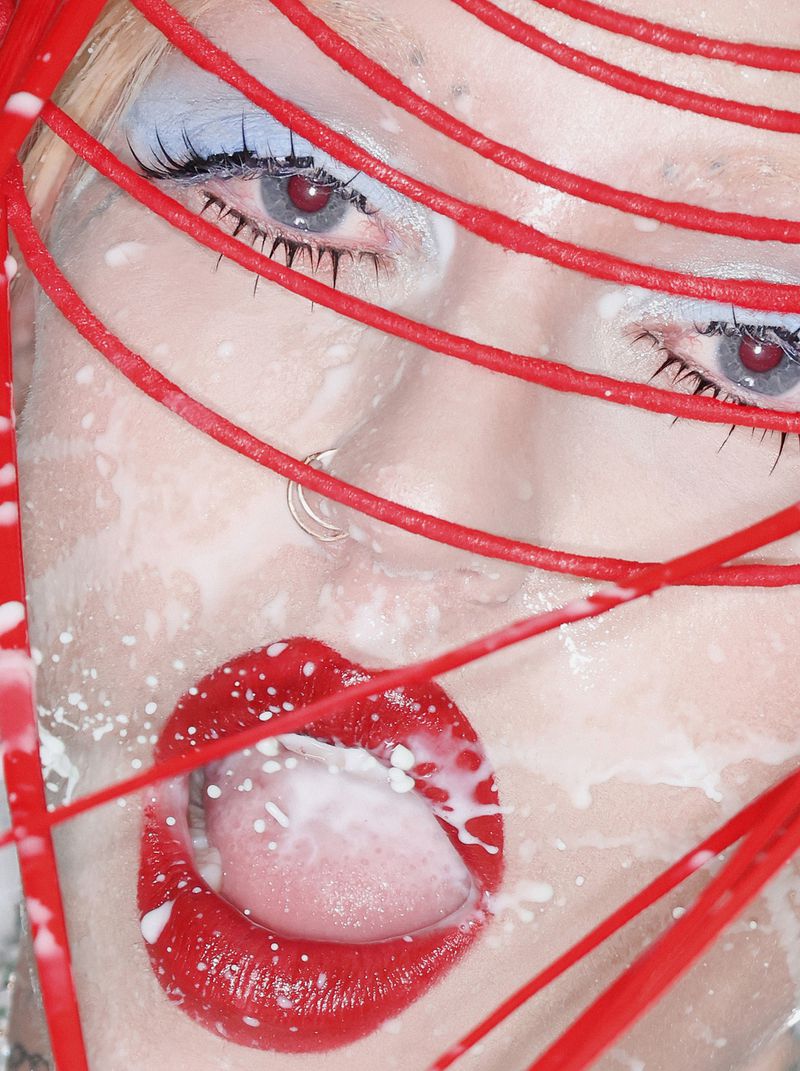 Artistic close-up portrait of a woman with striking red lipstick and makeup, featuring red cords and light liquid details.