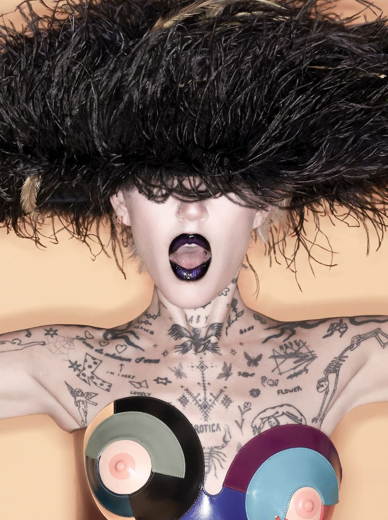 Portrait of a tattooed female with an open mouth wearing a large black feather headpiece and a colorful, abstract corset.