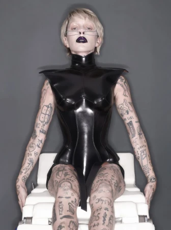 A woman with short blonde hair and tattoos sits on a white medical chair wearing a high-necked, dark-colored, latex bodysuit in a studio.