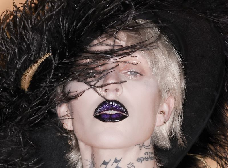 Dramatic beauty shot of a person with platinum hair and dark purple lips, wearing a feathered hat, photographed in a studio setting.