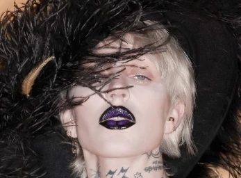 Brooke Candy wearing a large black feathered hat and glossy purple lipstick in a close-up portrait for The Perfect Magazine.