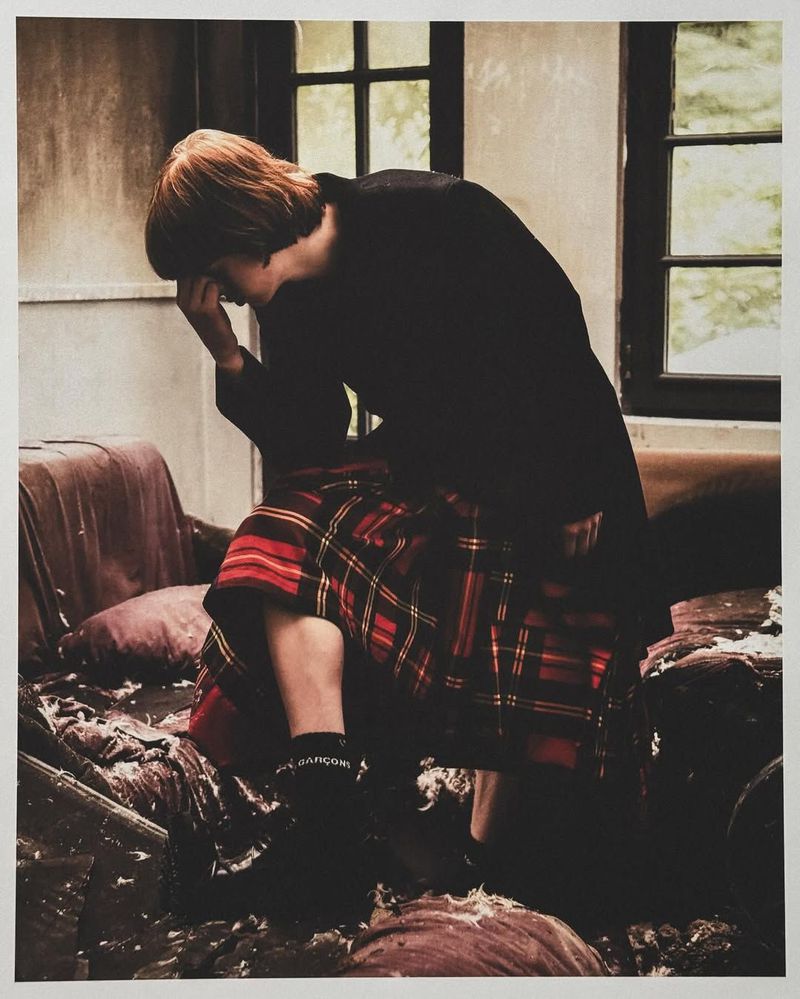 Person in a dark coat sitting on a damaged sofa, wearing a red and black plaid skirt, black socks, and black shoes, with a window behind.