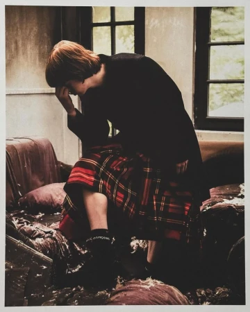 Person in a dark coat sitting on a damaged sofa, wearing a red and black plaid skirt, black socks, and black shoes, with a window behind.