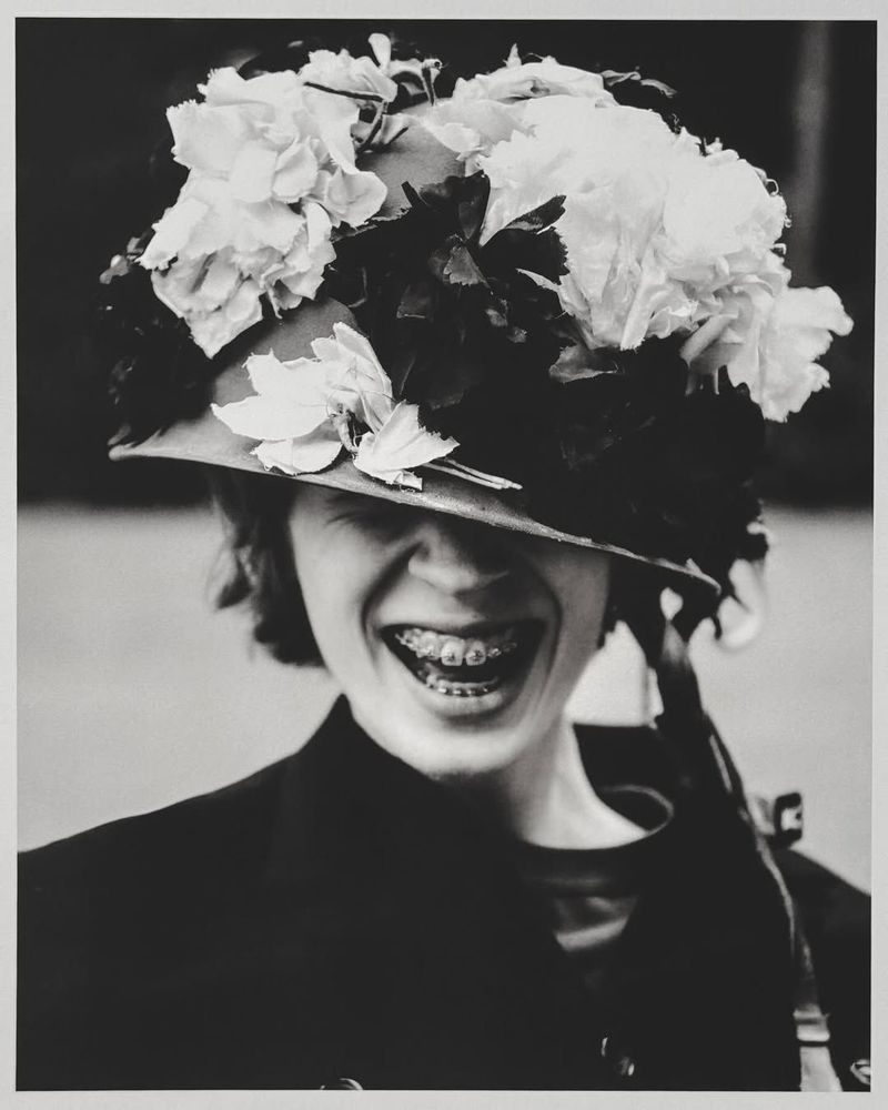 Black and white portrait of a smiling person wearing a floral-decorated military-style helmet, braces visible, against blurred backdrop.