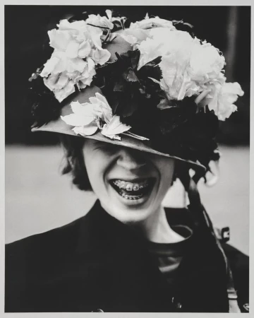 Black and white portrait of a smiling person wearing a floral-decorated military-style helmet, braces visible, against blurred backdrop.