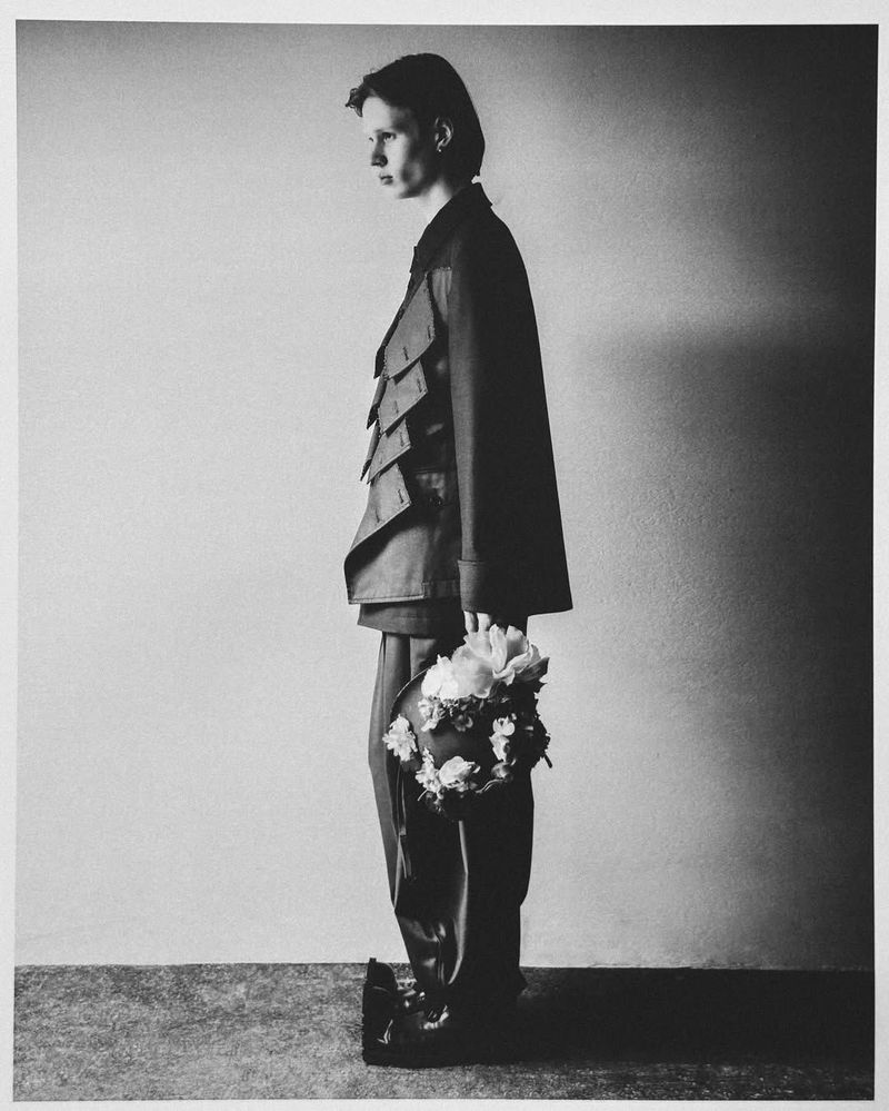 Monochrome portrait of a person in a sculptural jacket holding flowers, side view, in a simple indoor setting with soft lighting.
