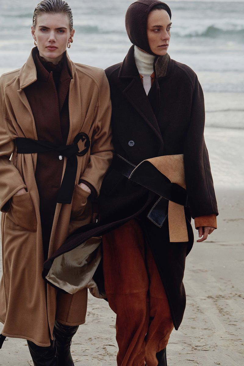 Two women in stylish winter coats walk on a sandy beach near ocean waves. Fashionable cold-weather clothing and neutral color palette.