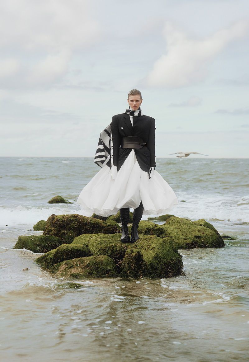 A woman in a black jacket and white skirt stands on mossy rocks by the sea under a cloudy sky, creating a striking fashion scene.