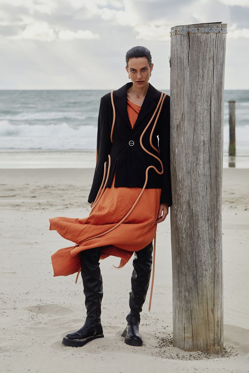 Model posing on a sandy beach beside a wooden post wearing a black blazer with cord trim, orange skirt, and black boots.