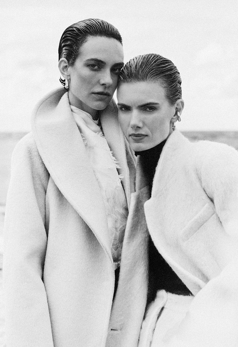 Two women pose in matching winter coats against a bright outdoor backdrop, showcasing an elegant, monochromatic fashion style.