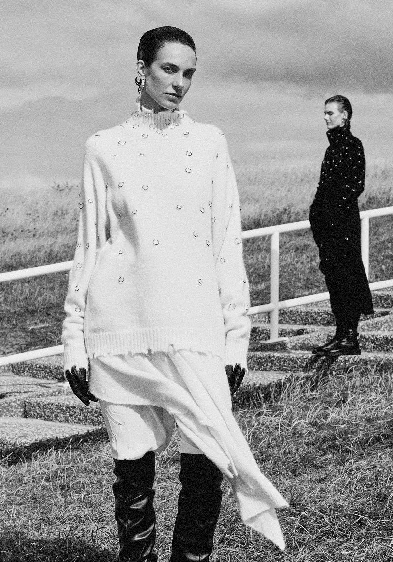 Black and white fashion portrait of a model in an embellished white knit sweater and tall black boots standing on a grassy path.